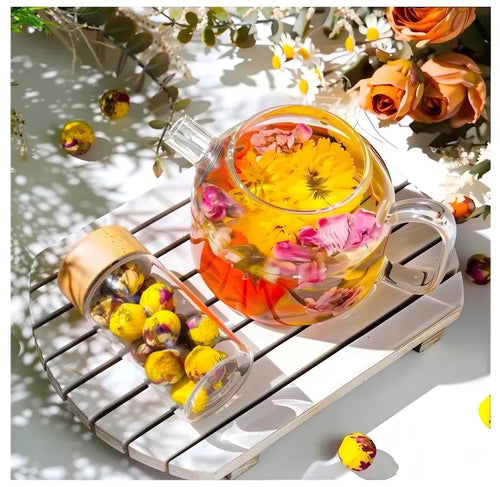 Clear glass teapot with floral tea on a wooden surface with flowers and leaves.